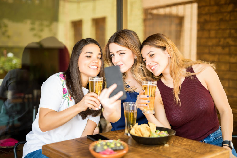 three young women sitting restaurant with beers taking selfie with mobile phone